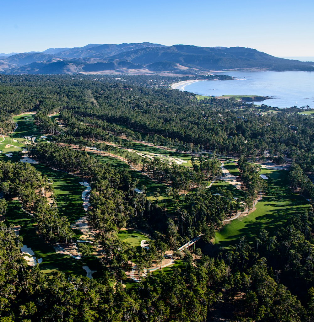 Aerial shot of Poppy Hills Golf Course with fairways and greens in the foreground and the Monterey Bay in the background.
